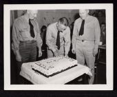 USS Saratoga birthday party, 3 men with middle one cutting a cake  
